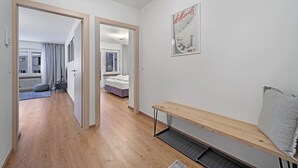 Un pasillo con suelo de madera que conduce a un dormitorio con cama y ventana. Hay un banco con cojín y un cartel en la pared.