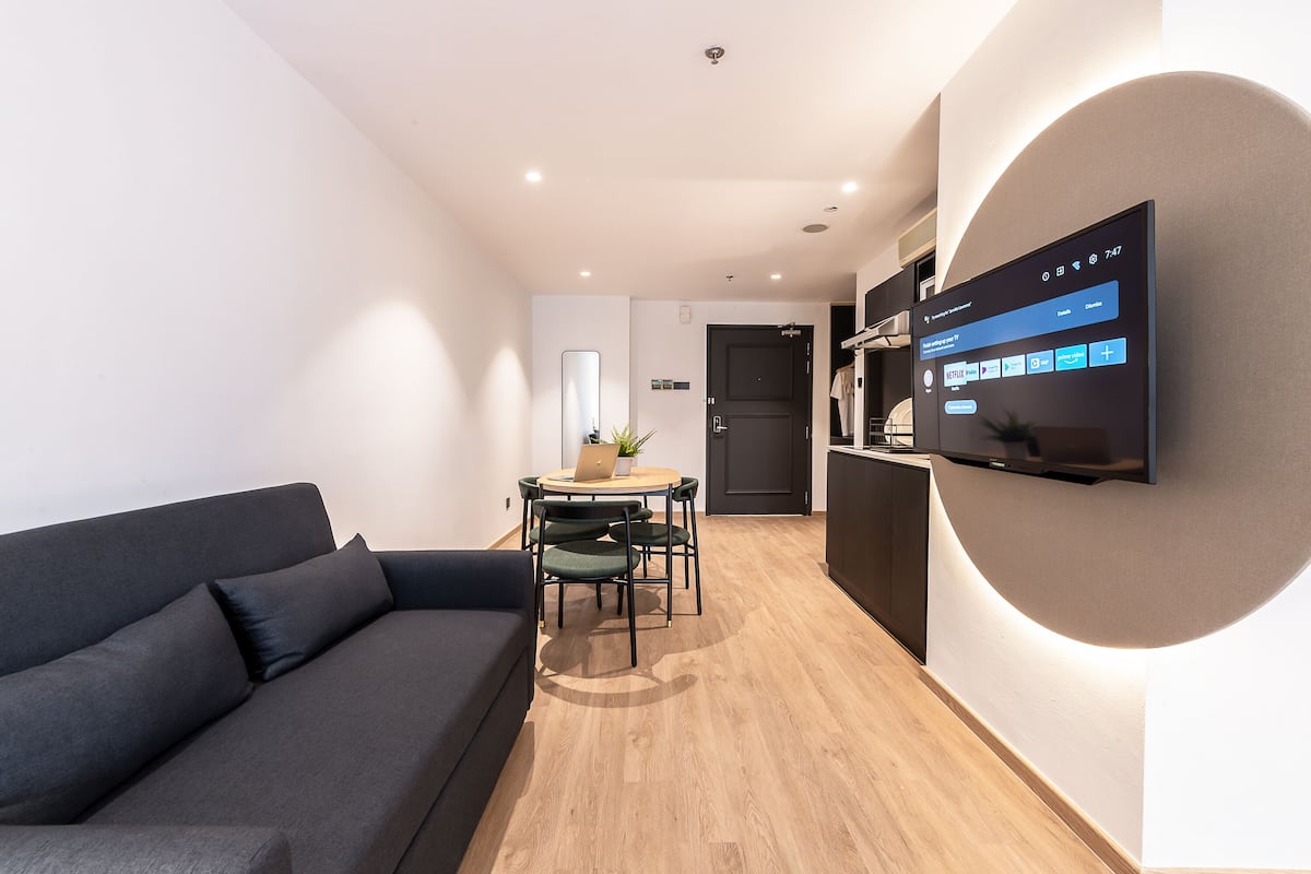 Een moderne woonkamer met een donkere bank, een houten eettafel met stoelen en een flatscreen televisie aan de muur.