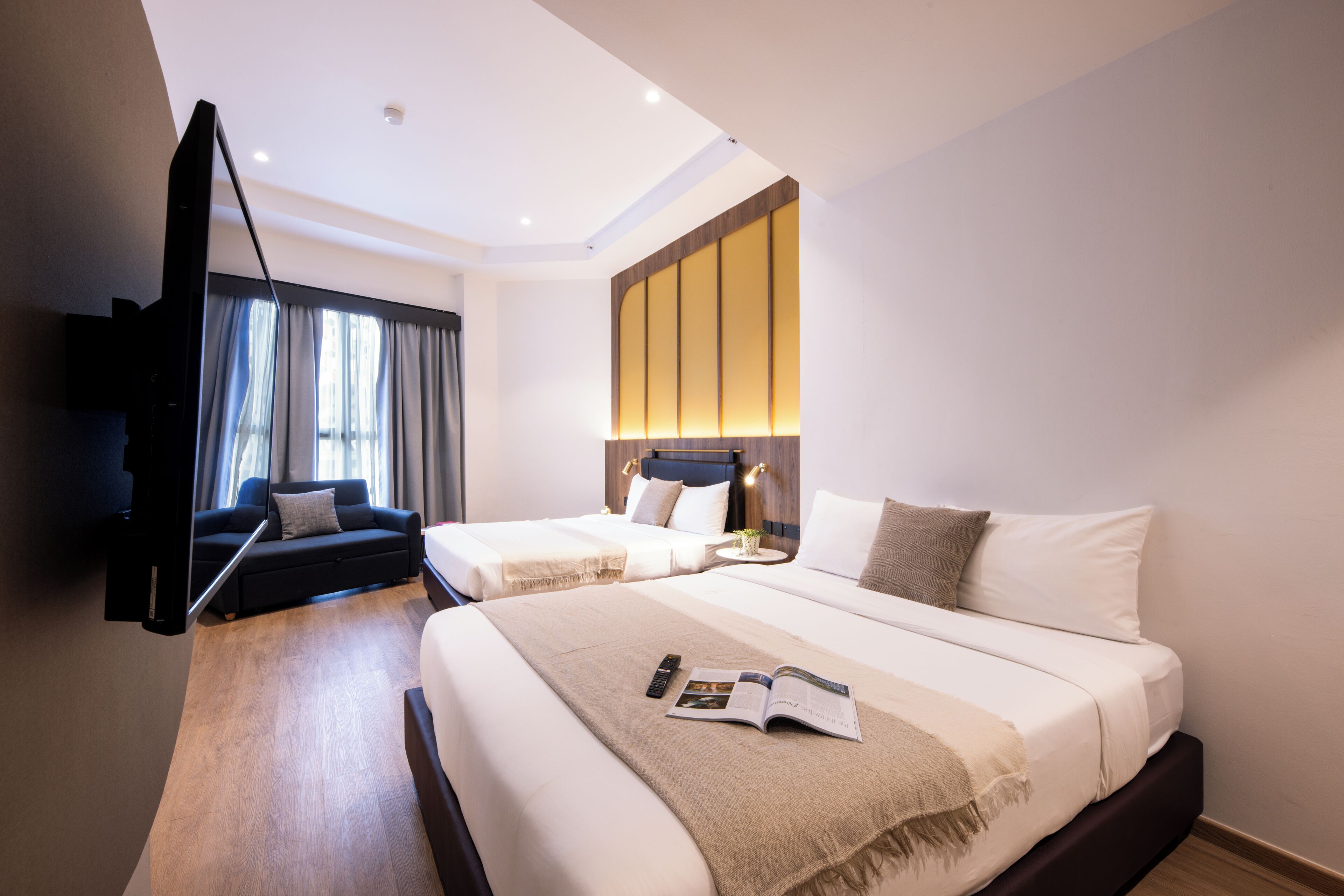 A hotel room with two beds, a sofa, a TV, and a bedside table with a lamp.