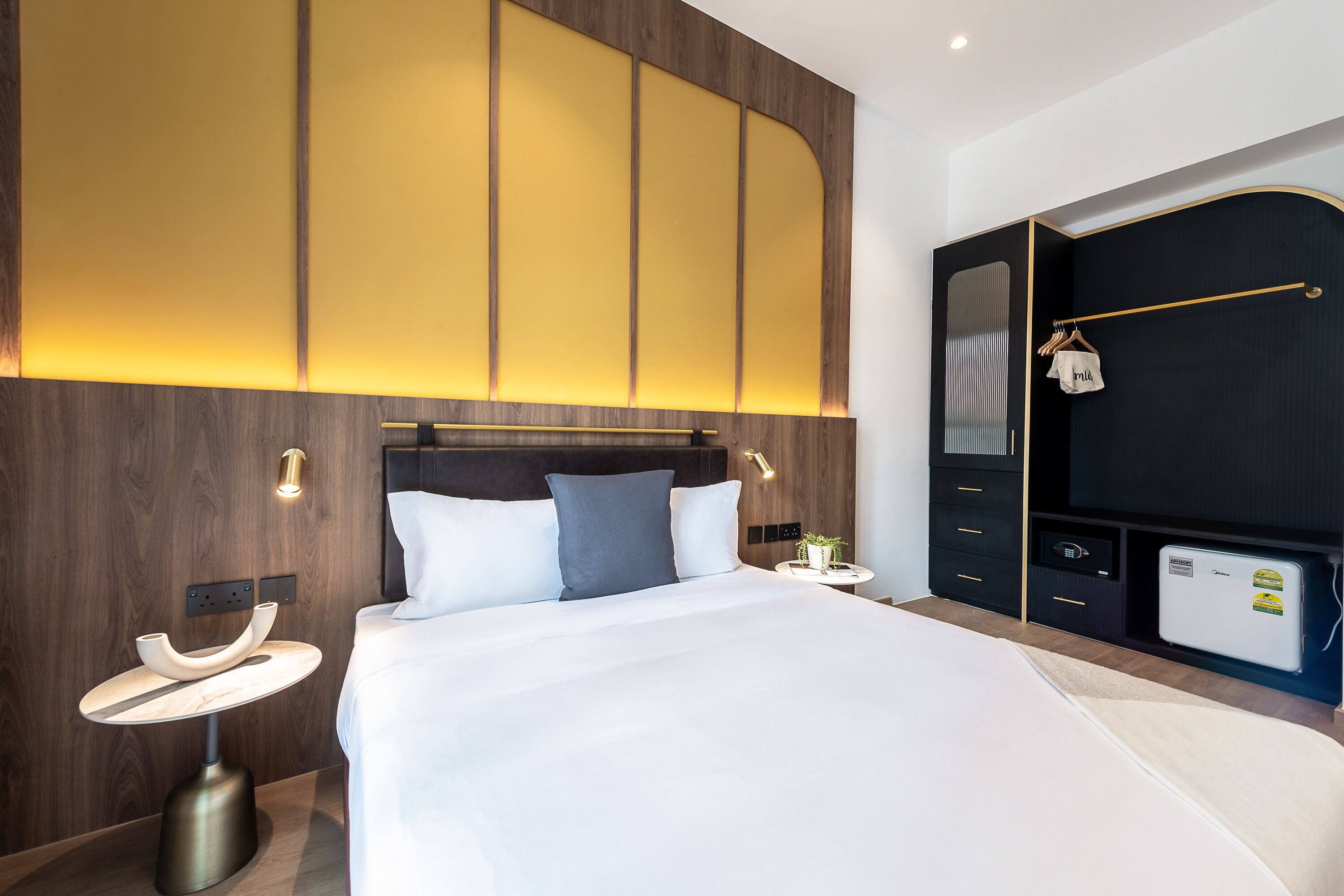 A modern hotel room with a large bed, bedside tables, a wardrobe, and a small refrigerator.