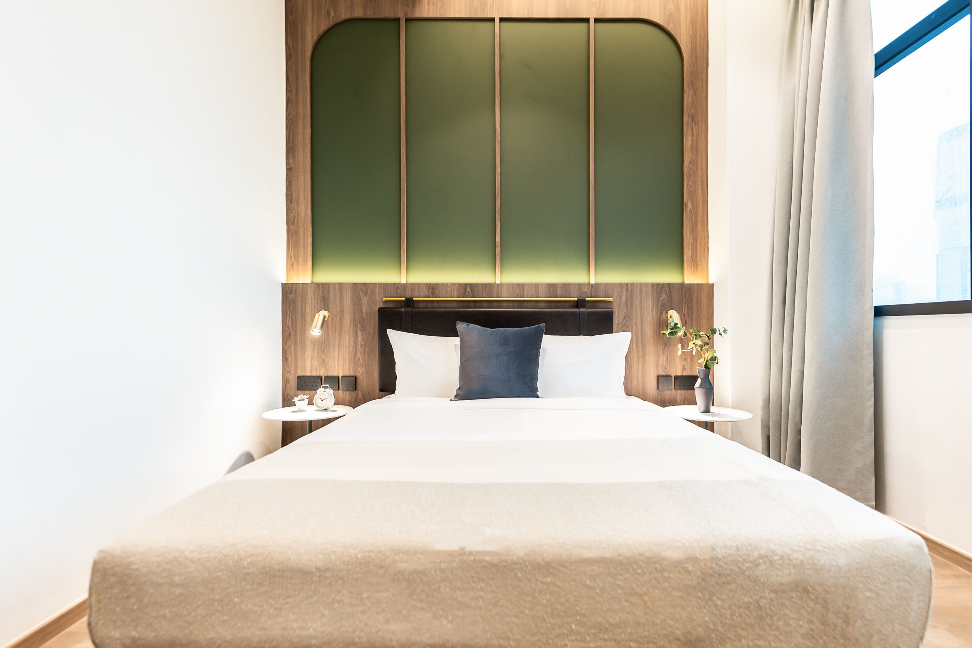 A modern hotel room with a large bed, wooden headboard, and a view of the city through a window.