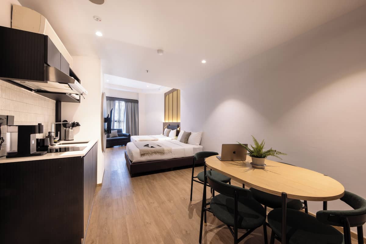 Een moderne hotelkamer met een kitchenette, een bed bij een groot raam, een eethoek met een tafel en stoelen, en een kleine potplant.