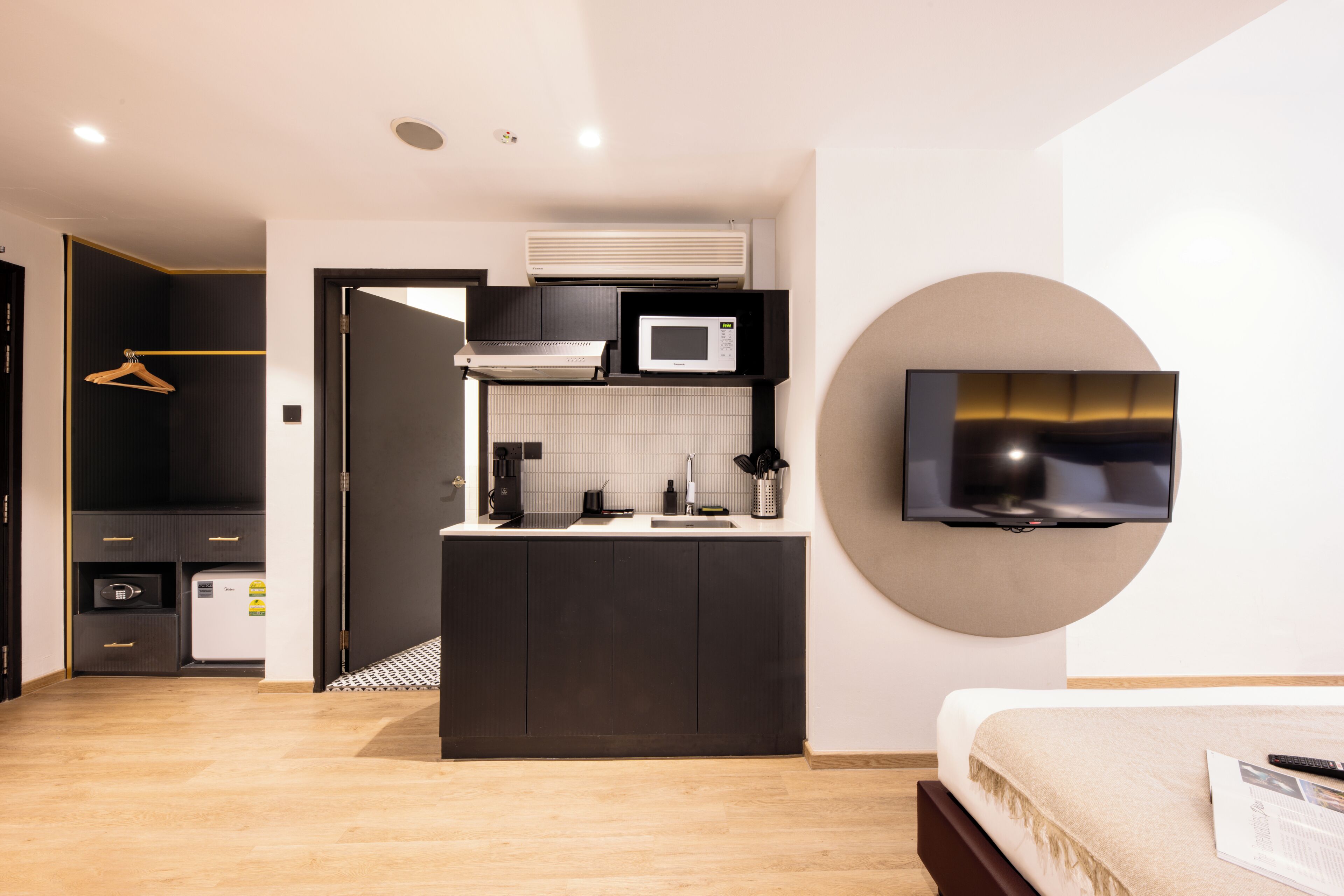 A modern hotel room with a kitchenette, a flat-screen TV mounted on the wall, and a bed with a remote control on it.