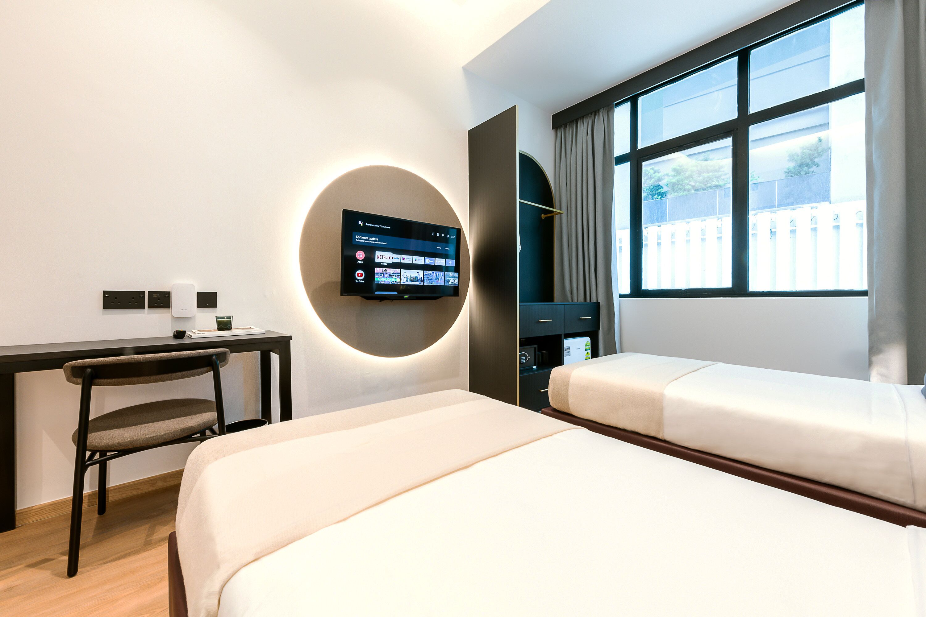 A hotel room with a bed, a desk, a chair, a TV mounted on the wall, and a window with curtains.
