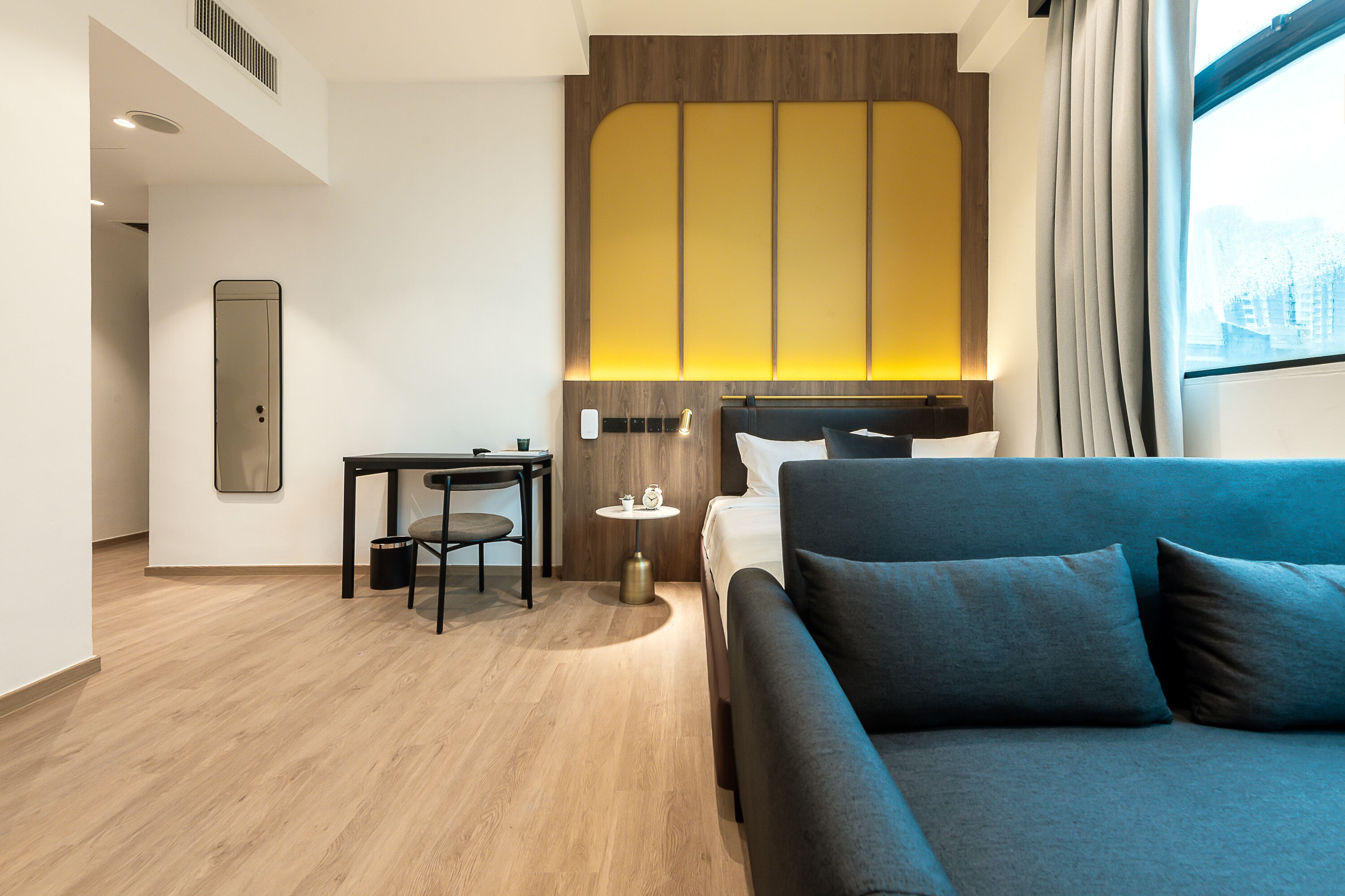 A modern hotel room with a wooden floor, a blue sofa, a small table with a chair, a bedside table with a lamp, and a large window with curtains.