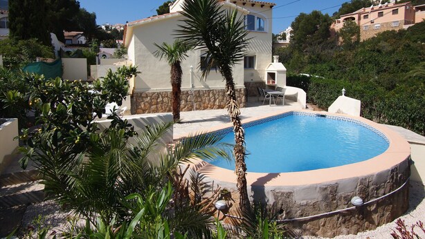 A pool area with a stone wall, palm trees, and a building with a tiled roof. - Charming detached holiday home with private pool–close to beach and fully fenced (Dénia)