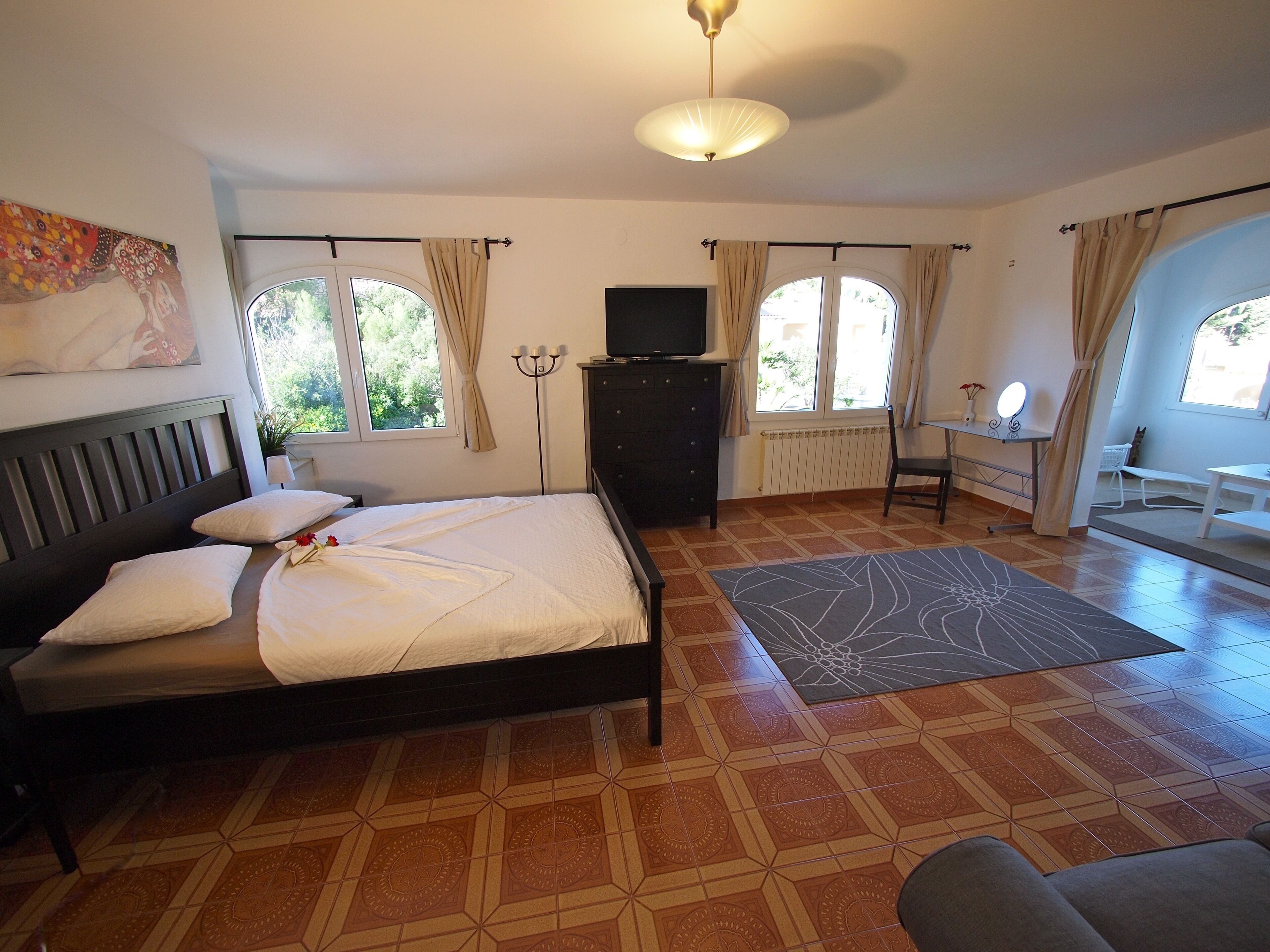 A bedroom with a bed, a dresser, a desk, and a chair. There are windows with curtains, a radiator, and a ceiling light.
