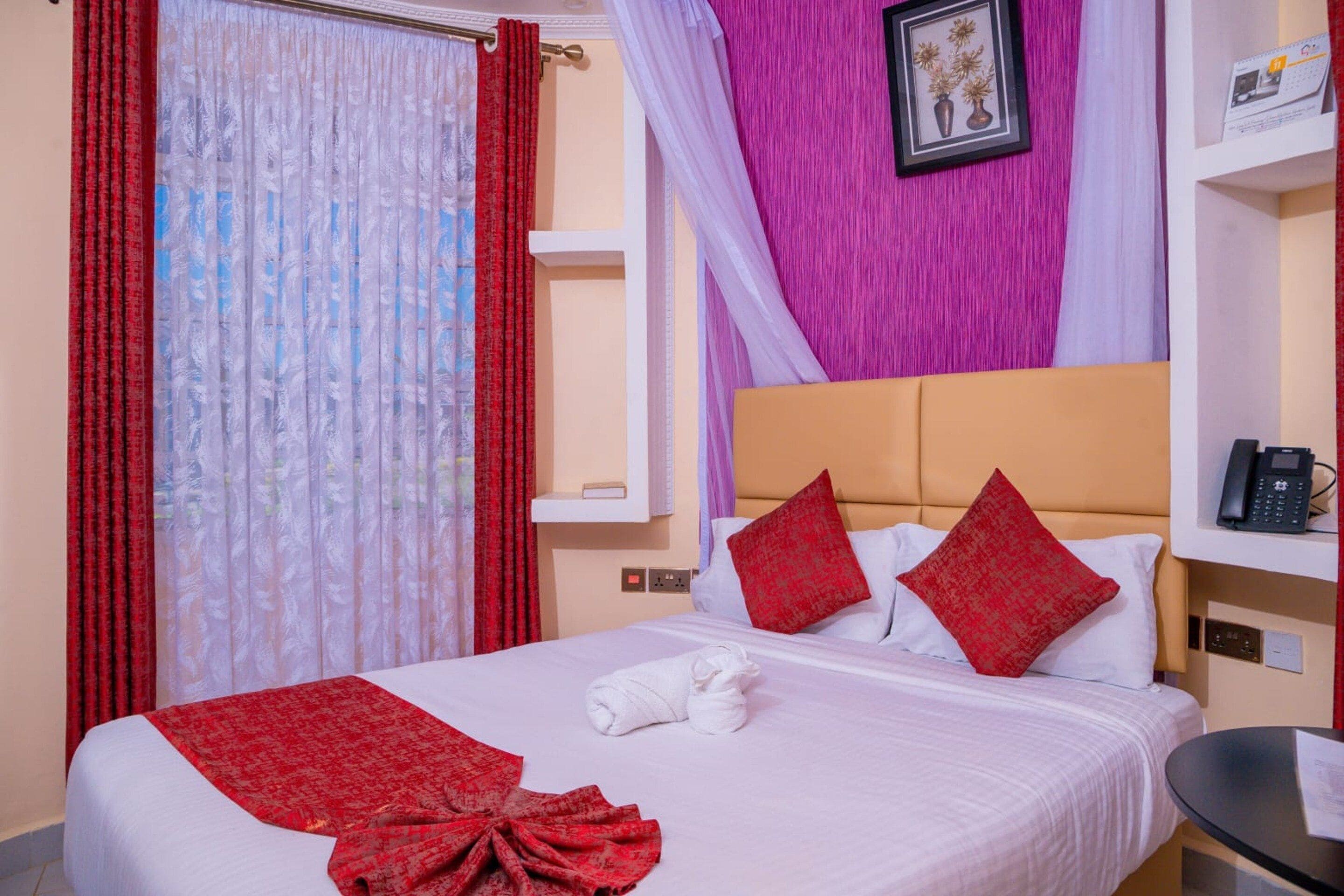 A hotel room with a bed, red and white curtains, a headboard, a bedside table with a phone, and a wall-mounted shelf with a framed picture.