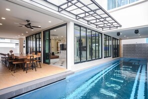 An indoor swimming pool with a glass enclosure, a ceiling fan, and a dining area with a table and chairs. - Exquisite Pool Villa H (Pattaya)