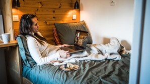 Una persona sentada en una cama con un ordenador portátil, un plato de comida y una taza sobre una mesita de noche.