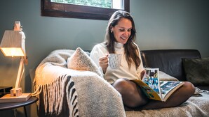 Una persona sentada en una cama con un libro y una taza, una lámpara, y una ventana al fondo.