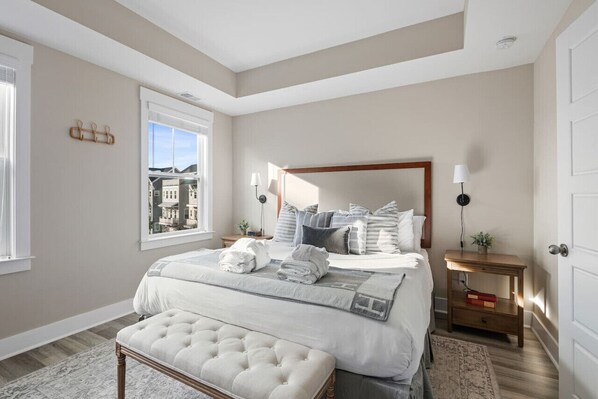Un dormitorio con una cama grande, una mesita de noche con lámpara, un banco y una ventana con vista a edificios.