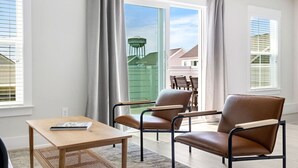 Ein modernes Wohnzimmer mit einem großen Schiebeglasfenster, das auf eine Terrasse führt, einem braunen Ledersessel, einem Holztisch und Blick auf ein grünes Gebäude.