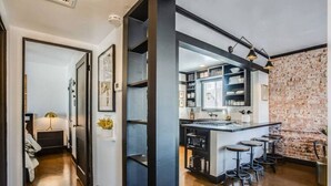 Een moderne keuken met een accentmuur van baksteen, een centraal kookeiland met barstoelen, en een gang met kunstwerken in lijsten.