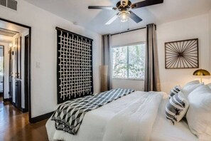 Une chambre à coucher comprenant un lit, un ventilateur de plafond, une fenêtre avec des rideaux et une œuvre d’art murale.