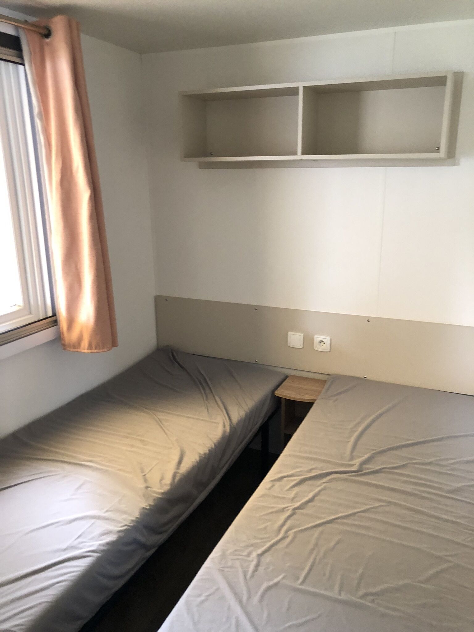 A small, single-bed room with a window, a wooden nightstand, and a shelf above the bed.