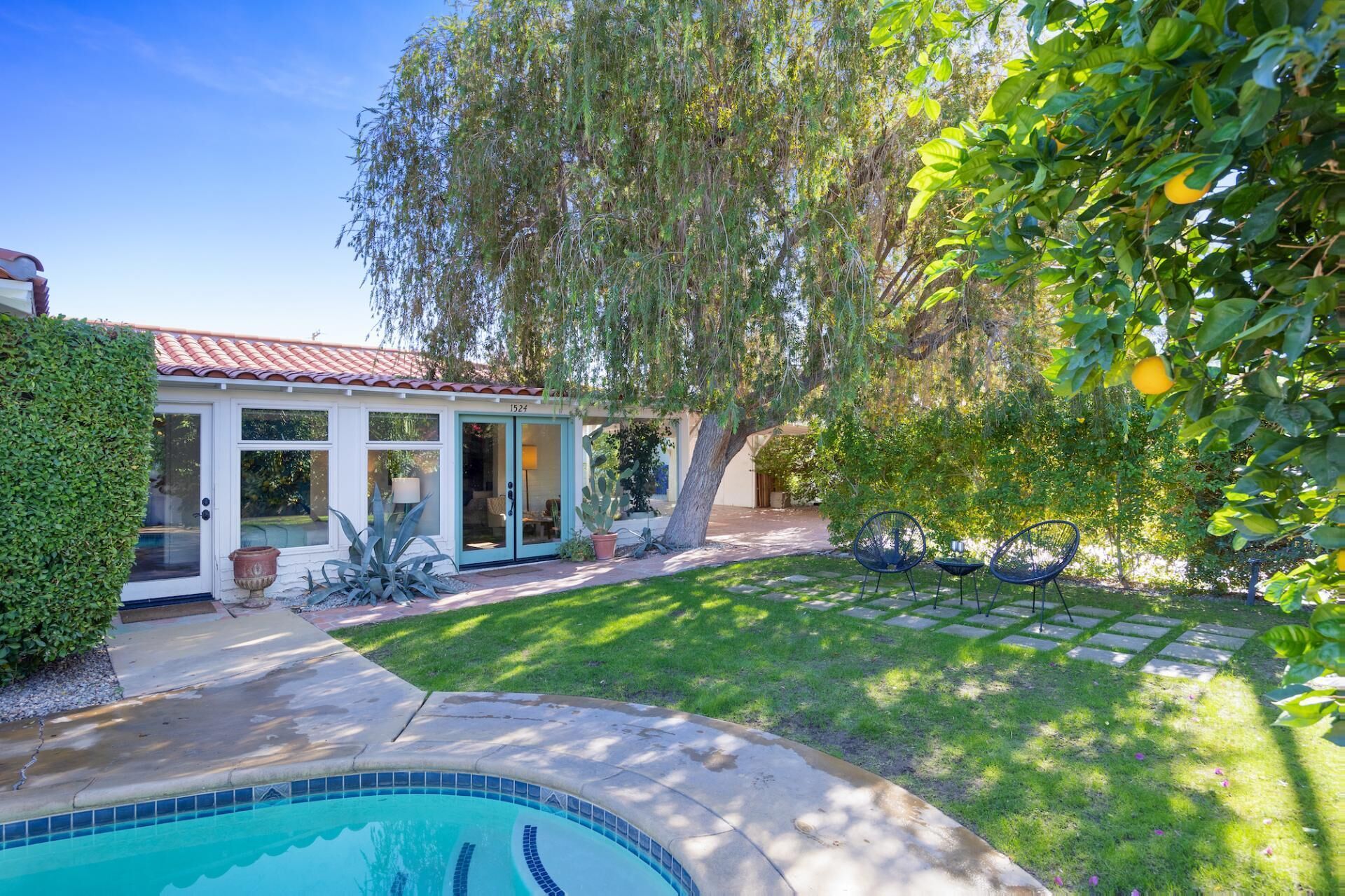 Una casa con piscina, un patio con sillas y un jardín con un árbol y un seto.