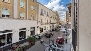 A narrow street with buildings on either side, featuring balconies, a row of parked cars, and outdoor seating areas. - 3141 B&b Incentro Comfort Suite - Central Suite by Barbarhouse (Lecce)