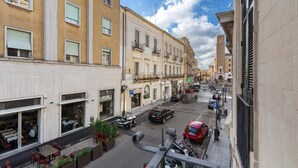 Una stretta via fiancheggiata da edifici, con balconi, una fila di auto parcheggiate e aree con tavoli all'aperto.
