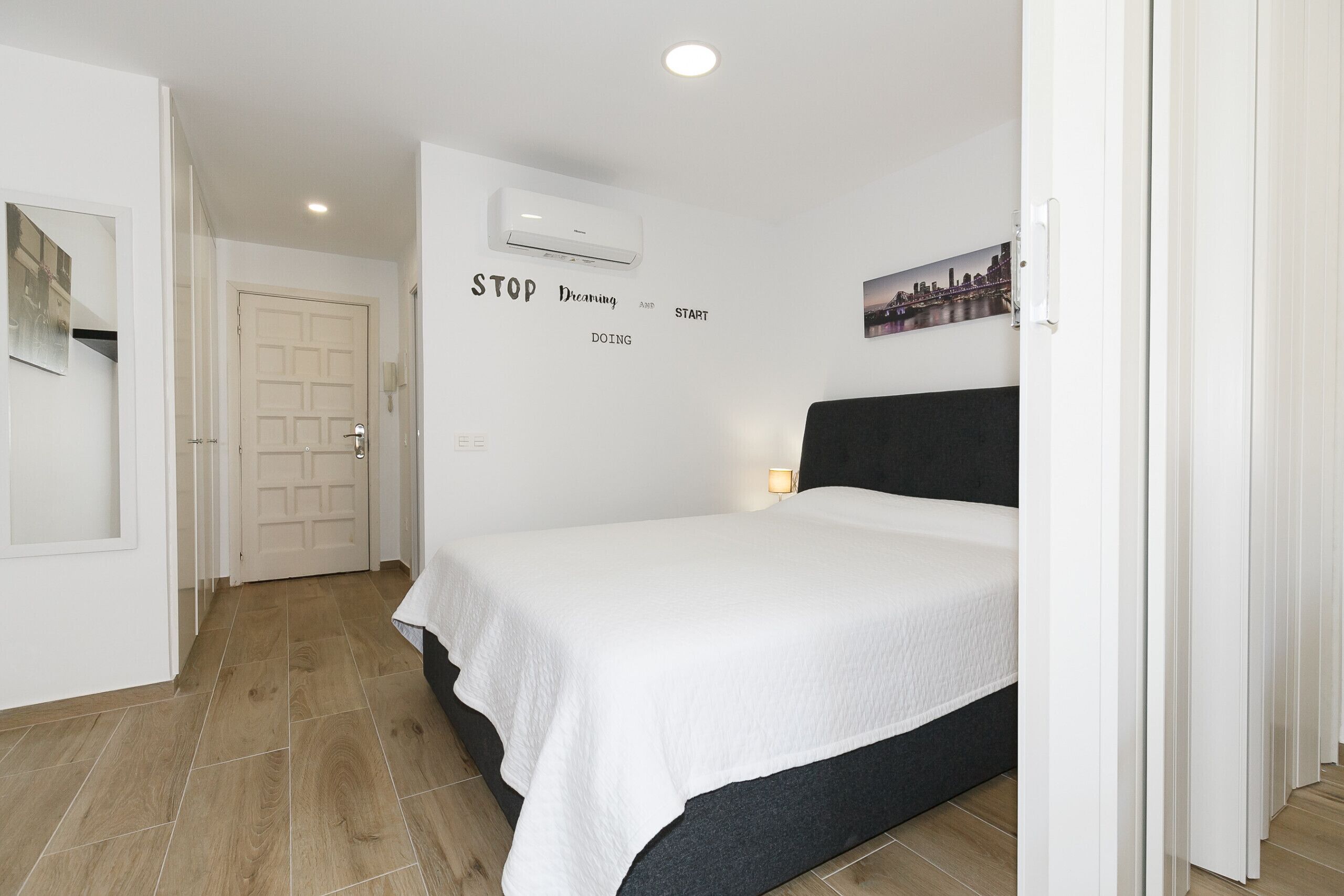 A bedroom with a bed, a wall-mounted air conditioner, a motivational quote, and a cityscape picture.
