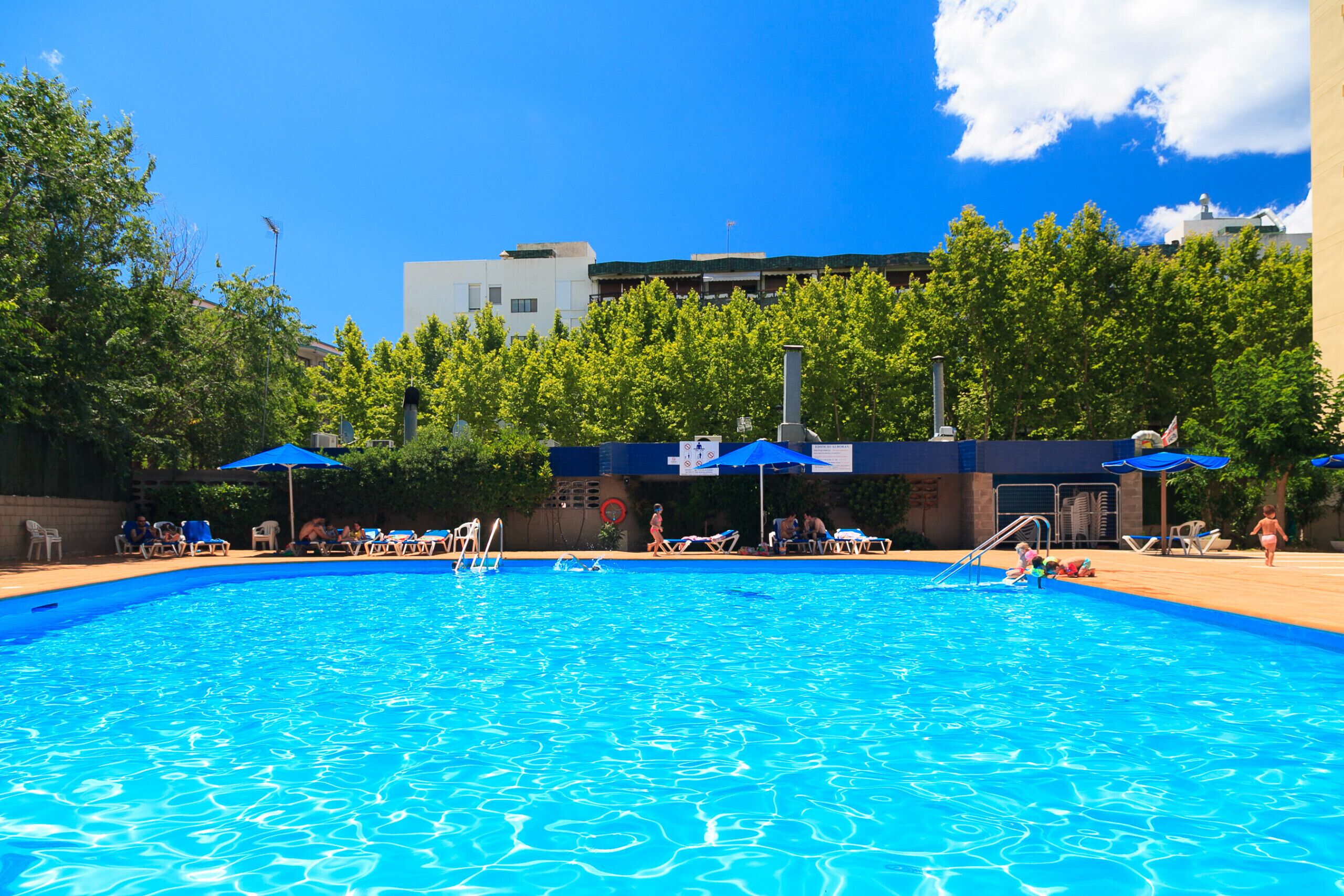 Ein Pool mit Liegestühlen und Sonnenschirmen, umgeben von Bäumen und Gebäuden.