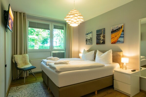 Una cama bien hecha con sábanas blancas, una lámpara de noche y un sillón con un cojín mullido.