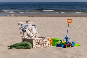 Escena de playa con una cesta de mimbre que contiene una toalla y una botella, una toalla verde y un tractor de juguete en la arena.