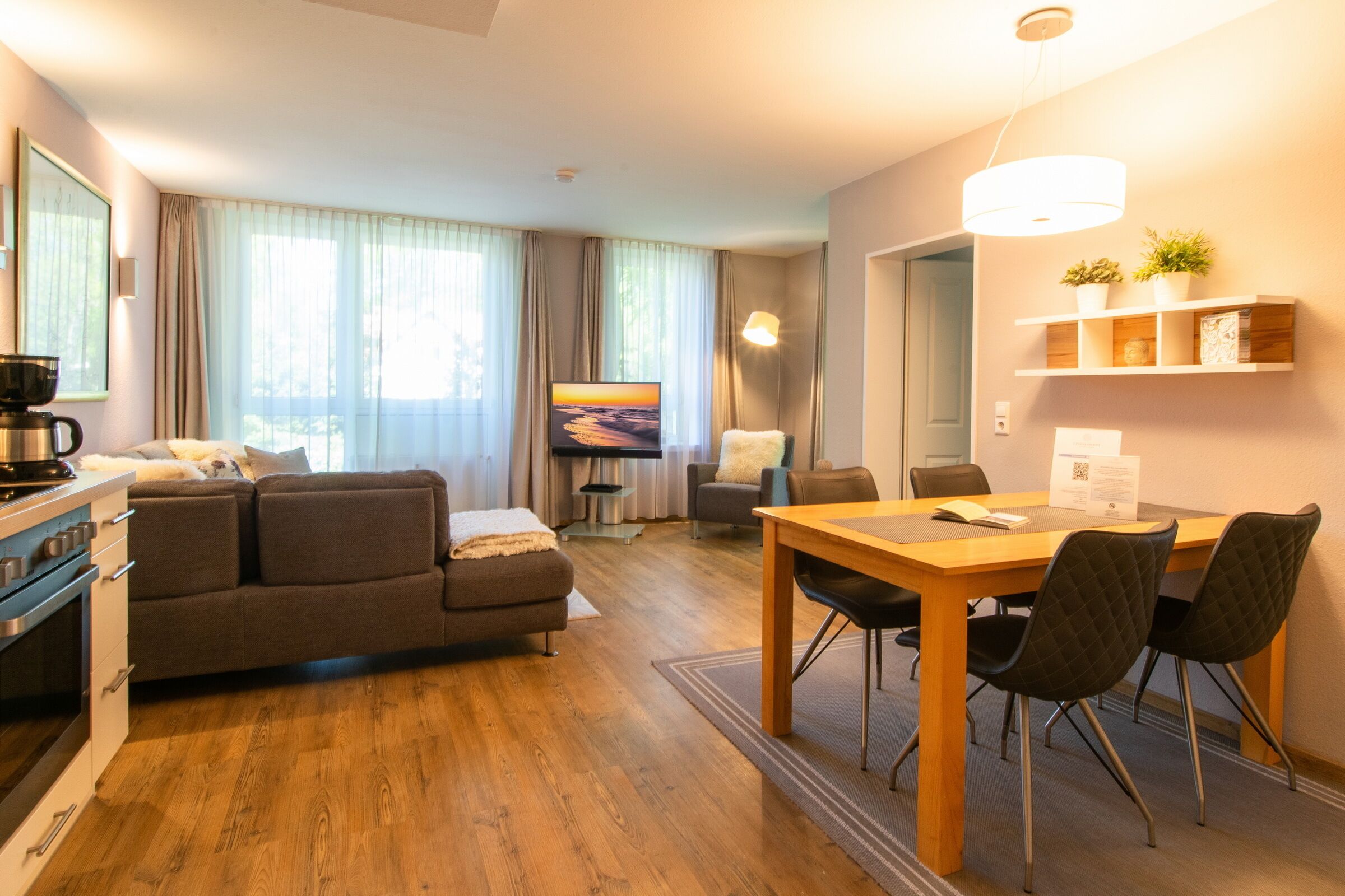 Moderni hotellihuone, jossa on puulattia, sohva, ruokapöytä tuoleineen, televisio ja kahvinkeitin.