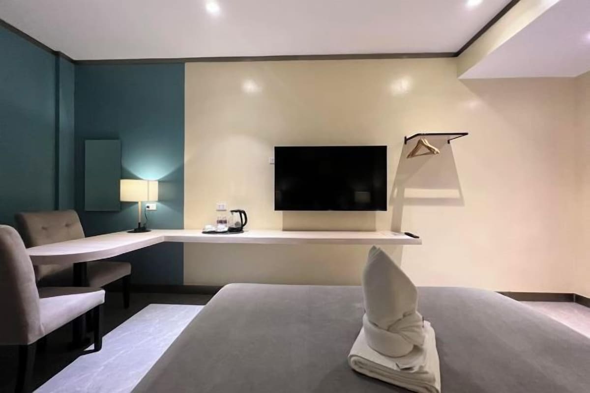 A hotel room with a bed, a desk, a chair, a flat-screen TV, a lamp, and a coat rack.