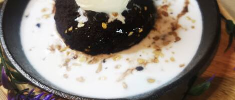 A dessert with a scoop of ice cream on top of a chocolate cake, served in a black bowl on a wooden board with purple flowers.