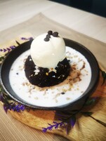 Postre con una bola de helado sobre un pastel de chocolate, servido en un cuenco negro sobre una tabla de madera con flores moradas.