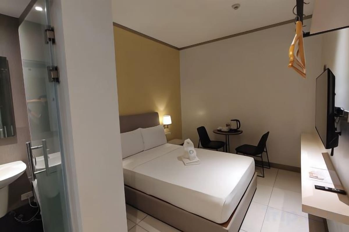 A hotel room with a bed, a desk, a chair, a small table, a TV, and a bathroom.