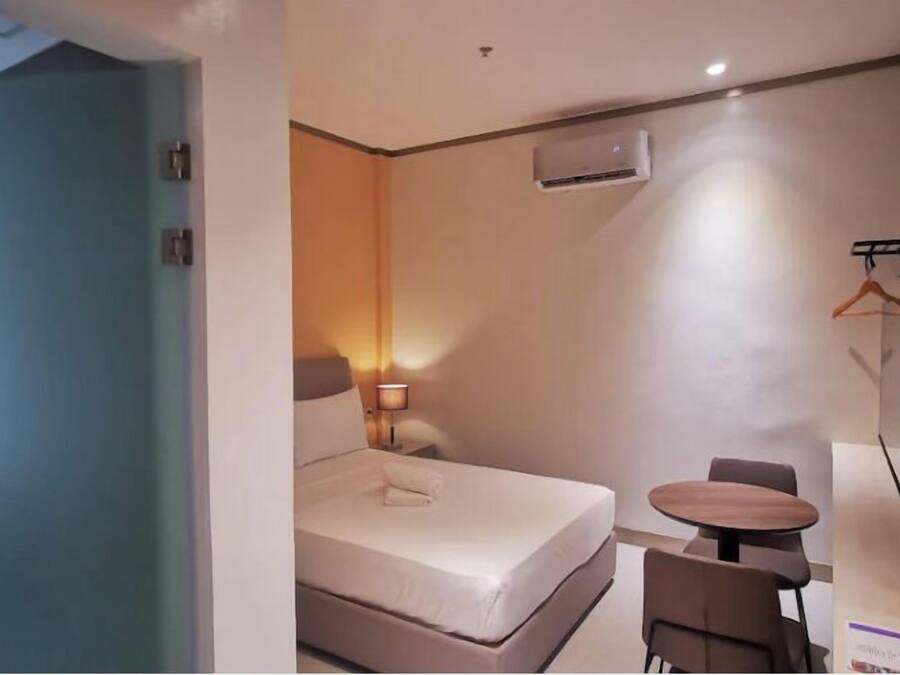 Habitación de hotel con cama, mesita de noche, silla y una pequeña mesa. Se observa un equipo de aire acondicionado en la pared y un perchero.