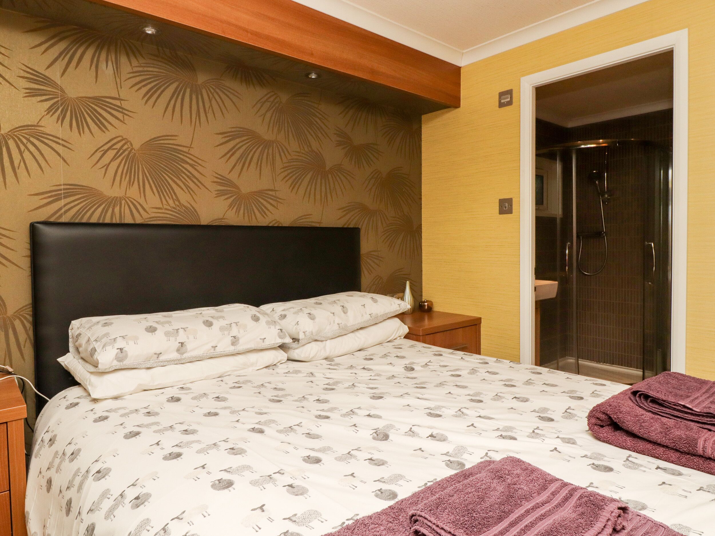 A bedroom with a bed, a nightstand, a wall with a palm leaf pattern, and a door leading to a bathroom.