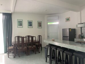 A modern kitchen with a dining area, featuring a marble countertop, wooden chairs, a refrigerator, and a window with curtains. - Remarkable Studio in Jungle of Chemuyil, Tulum (Tulum)