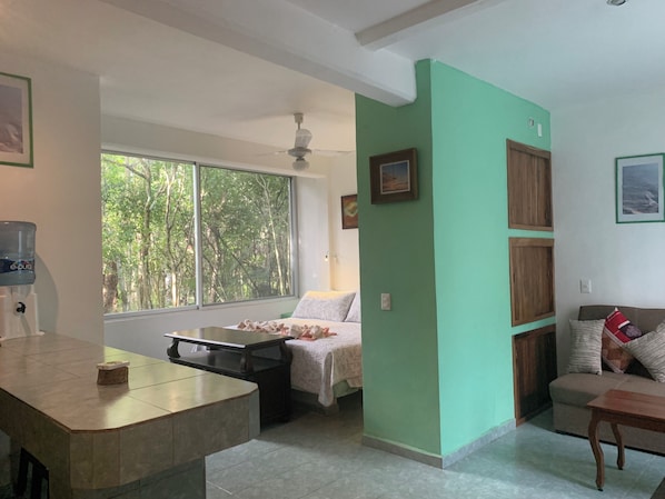 A room with a bed, a desk, a water dispenser, a window with a view of trees, and a ceiling fan. - Remarkable Studio in Jungle of Chemuyil, Tulum (Tulum)