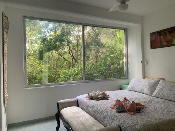 A bedroom with a large window, a bed with a patterned bedspread, a bench, and a painting on the wall. - Remarkable Studio in Jungle of Chemuyil, Tulum (Tulum)