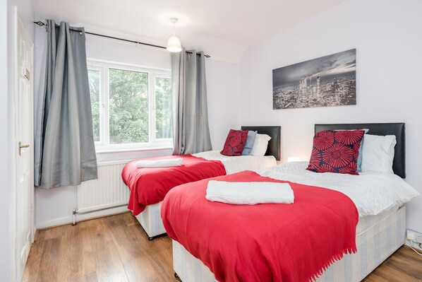 Hotellihuone, jossa on kaksi sänkyä, harmaat verhot, kaupunkimaisemaa esittävä taulu ja ikkuna, josta avautuu näkymä puita kohti.