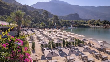 A beach resort with rows of sun loungers and umbrellas, a pool, and mountains in the background.