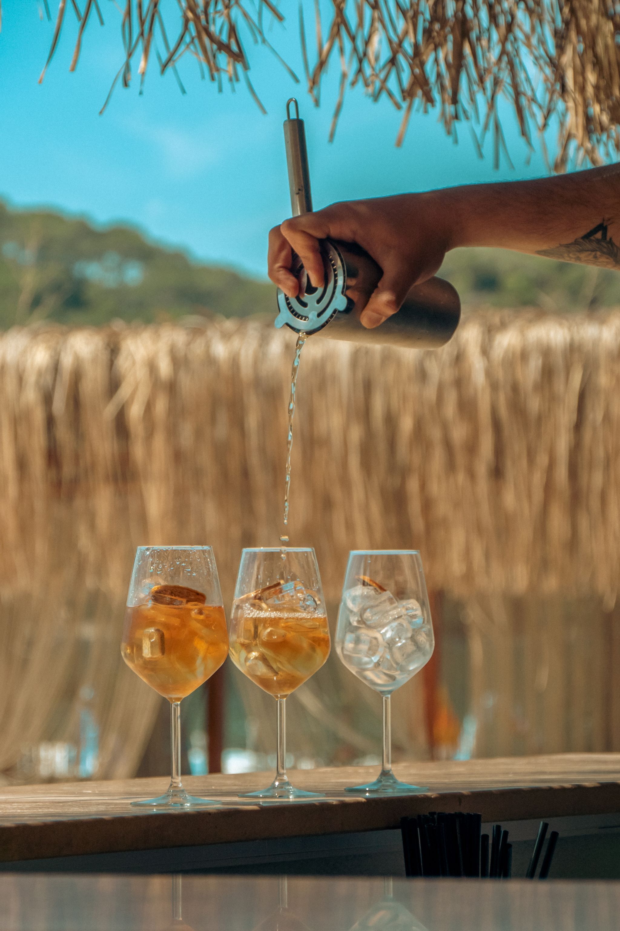 En hand häller upp en drink i tre glas på en bardisk, och ett sugrör ligger bredvid. Bakgrunden är suddig och visar ett naturligt landskap.