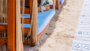 Une rangée de cabanes de plage en bois, dotées de toits de chaume et de rideaux, chacune comprenant un banc rembourré et une petite table.