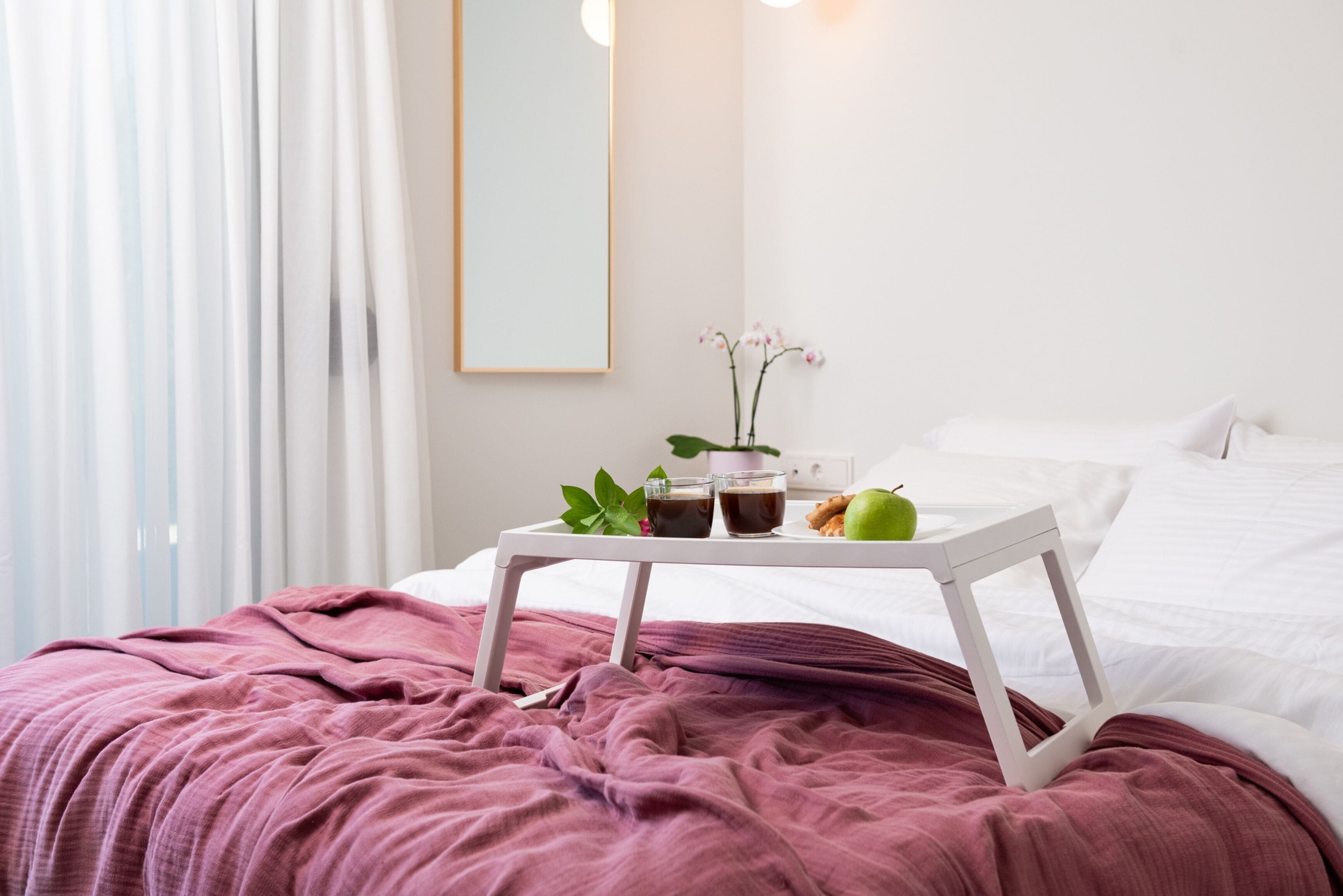 A neatly made bed with a white frame, a pink blanket, a small white table with a tray of food and drinks, and a potted orchid on the bedside table.