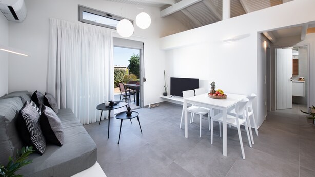 A modern living space with a grey sofa, a dining table with chairs, a flat-screen TV, and a small kitchen area.