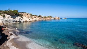 A coastal beach with clear water, rocky cliffs, and a person standing on the shore.