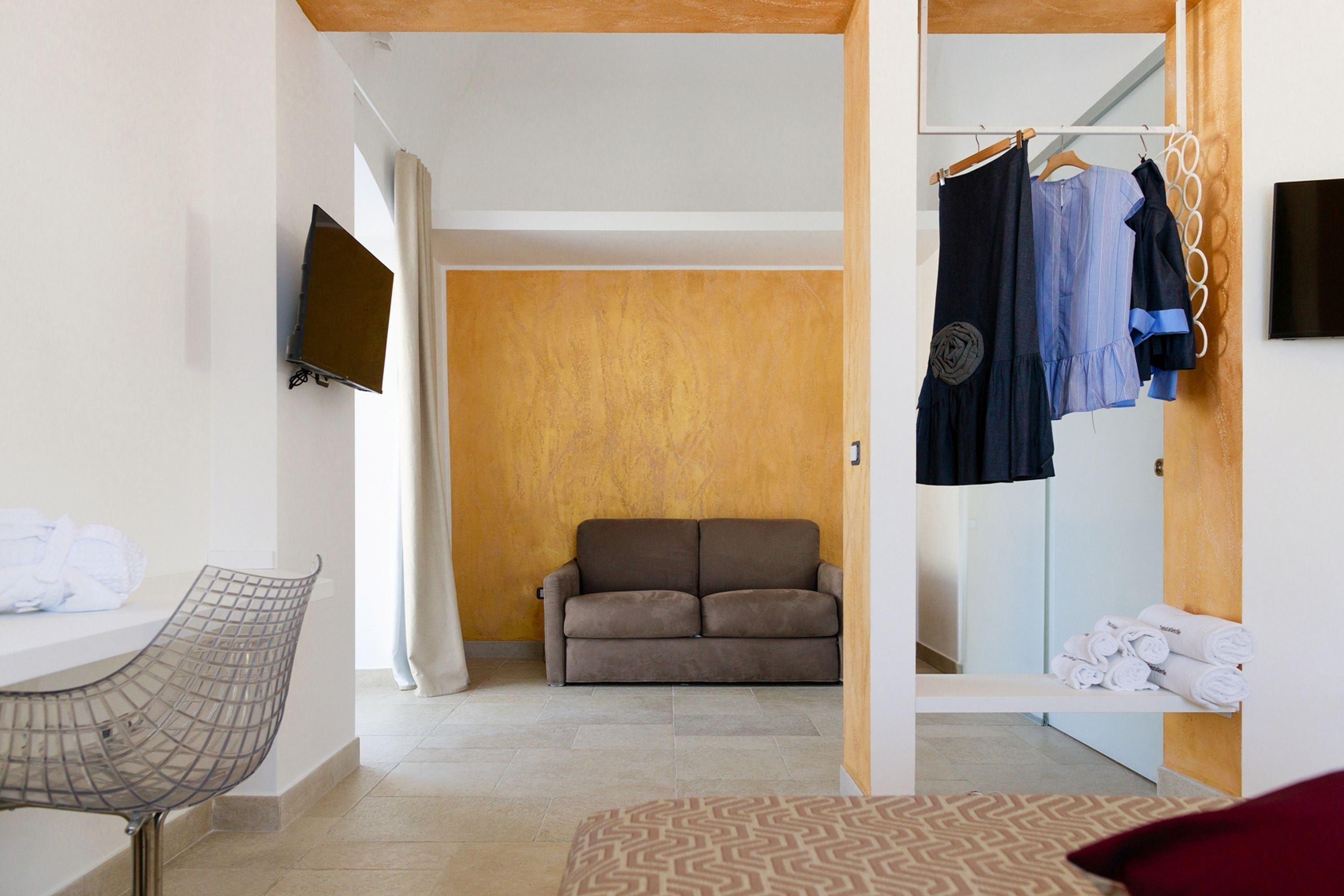 A hotel room with a sofa, a TV mounted on the wall, a chair, and a bed with patterned bedding.