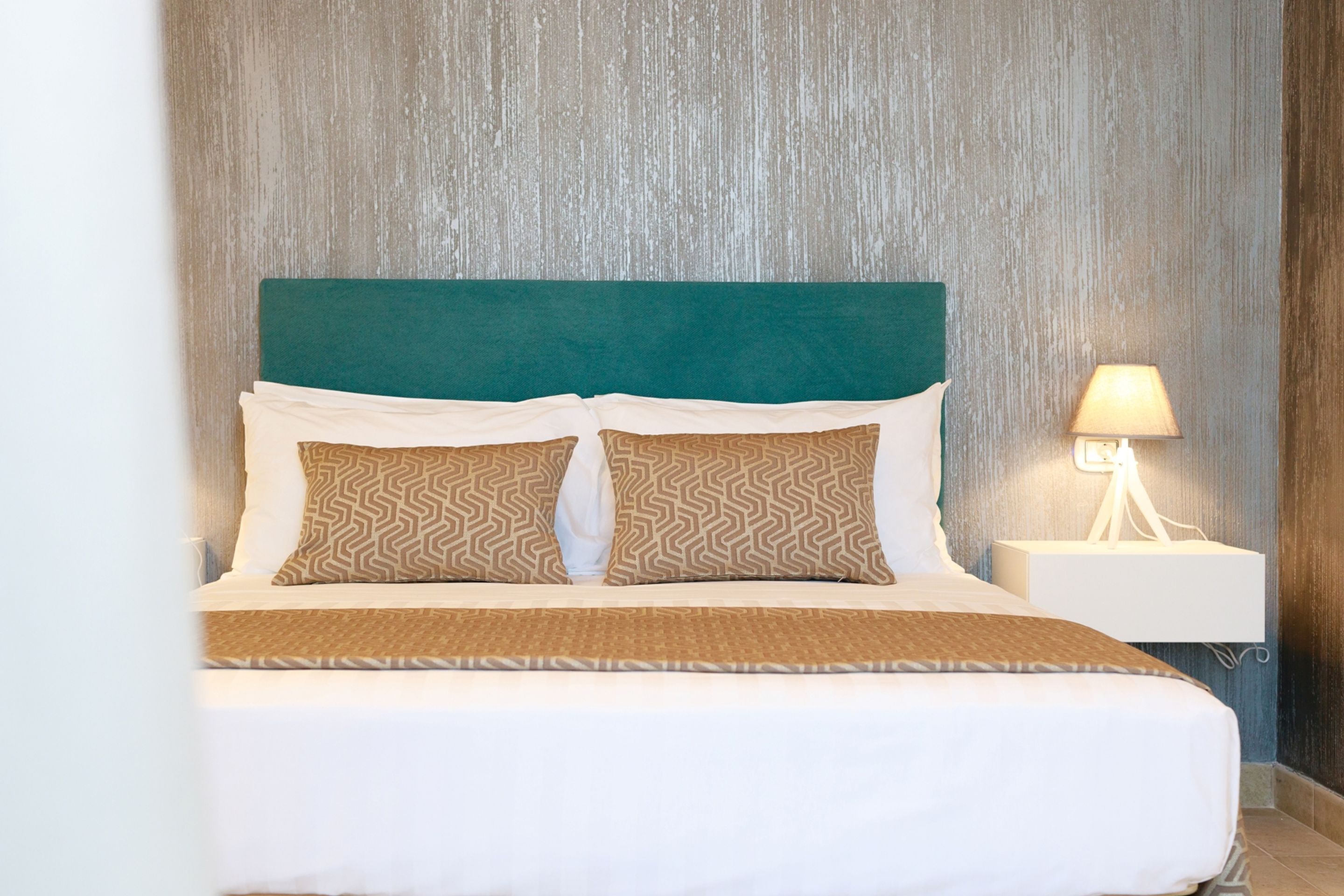 A neatly made bed with a green headboard, two patterned pillows, and a white bedside table with a lamp.