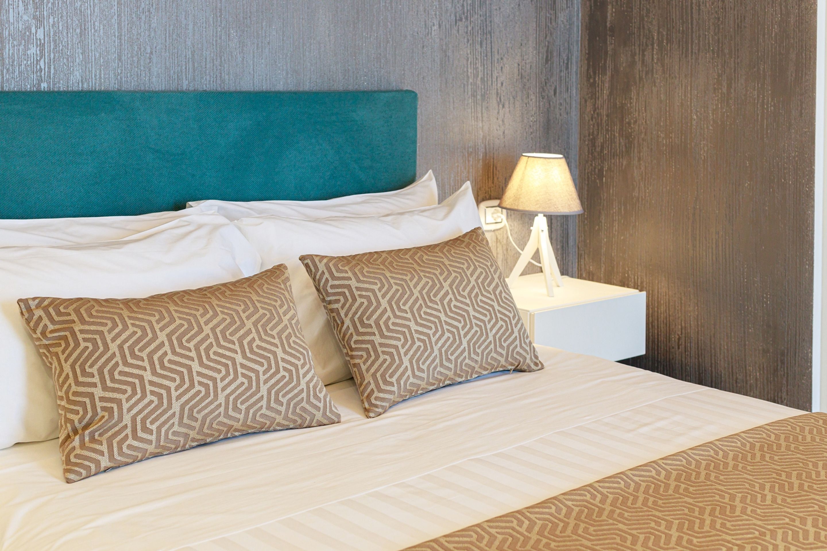 A neatly made bed with patterned pillows, a teal headboard, and a bedside lamp.