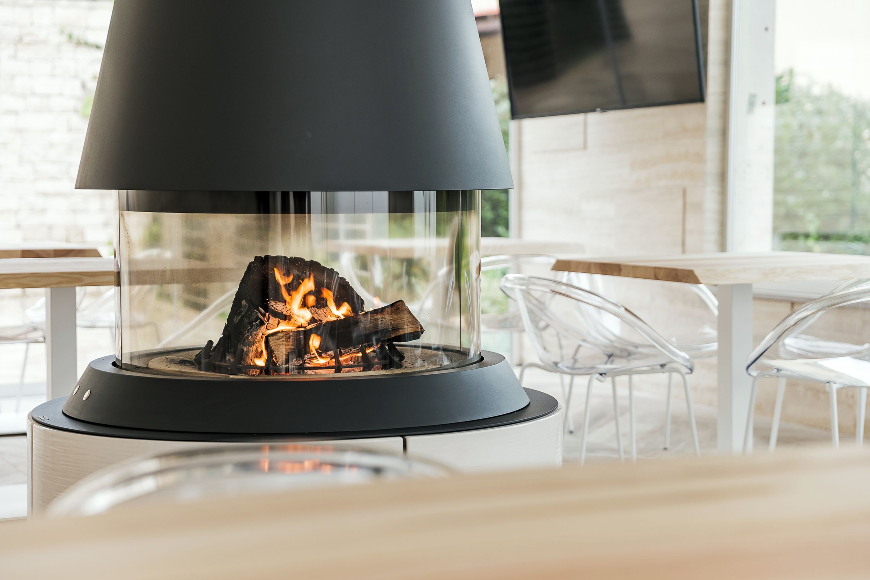 A modern outdoor seating area with a glass-enclosed fire feature, clear chairs, and a wooden table.