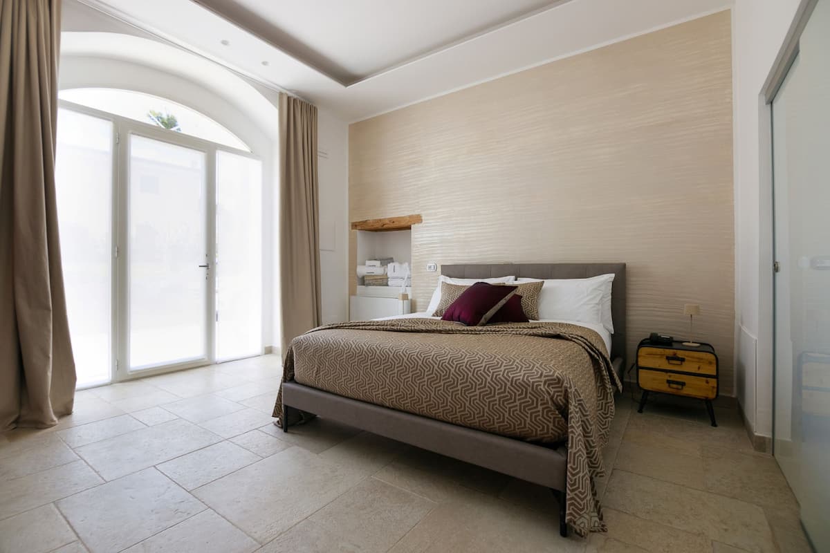A bedroom with a large bed, a nightstand, a wooden dresser, and a sliding glass door leading to a balcony.