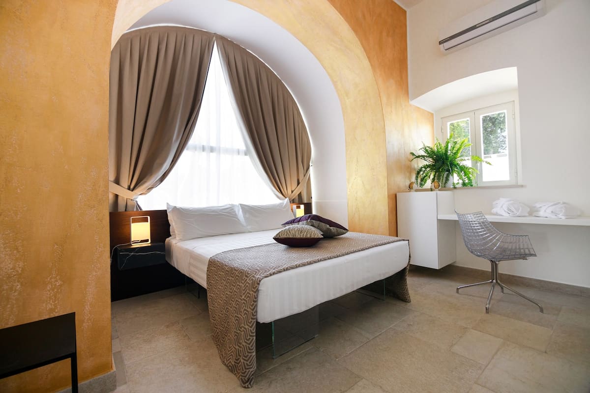 A bedroom with a bed, curtains, a desk, a chair, and a potted plant.